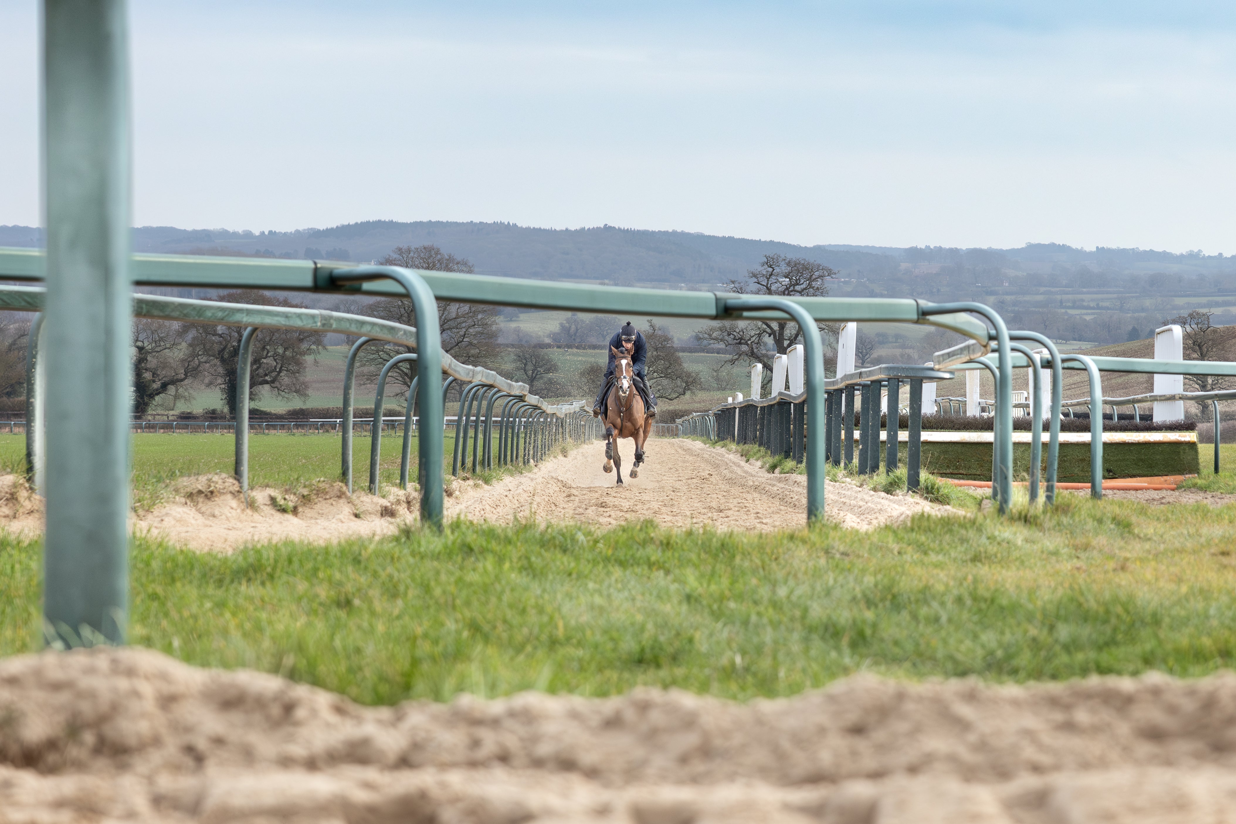 Jockey Harry Skelton riding a galloping horse on a sandy racetrack, framed by green training rails.