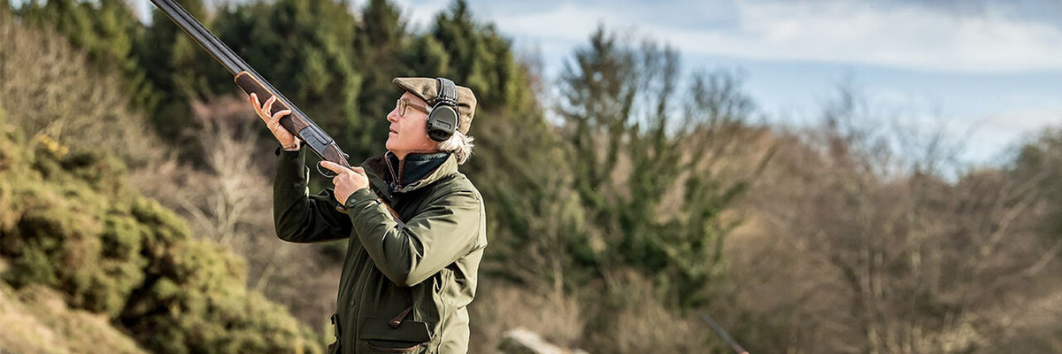 Man in olive green jacket aims double-barreled shotgun for partridge hunting