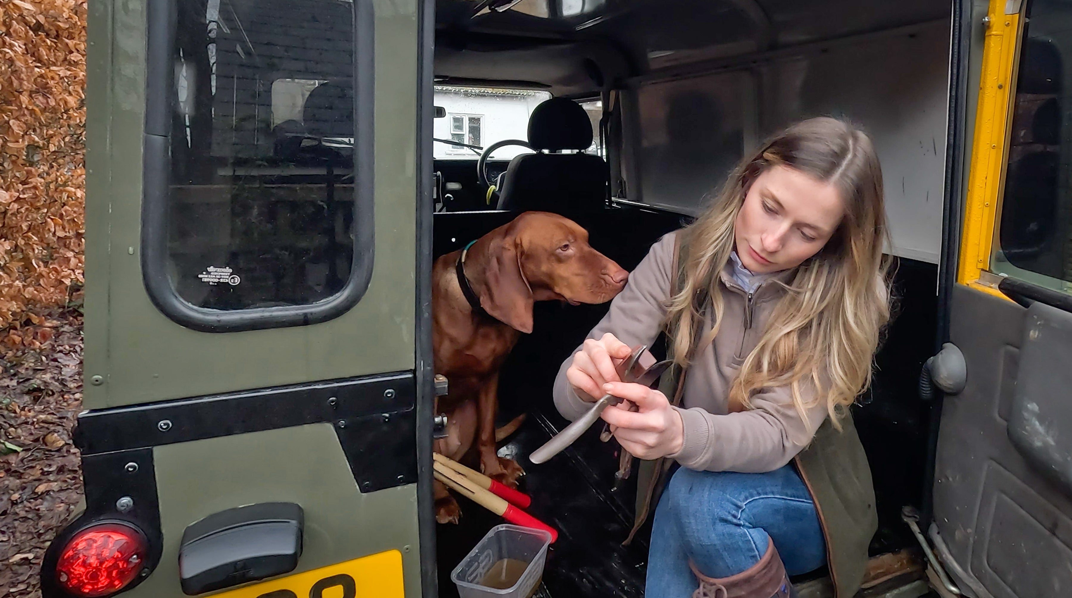 Woman with long blonde hair examines gray dog collar in green Land Rover