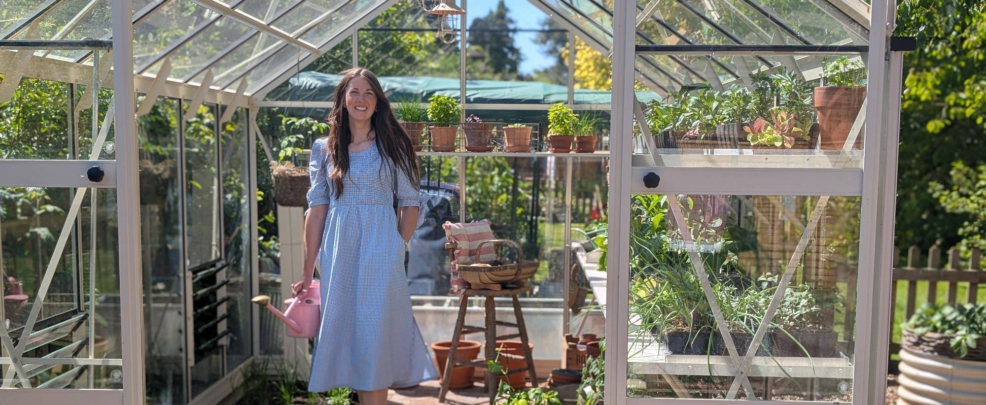 Amy Shore in light blue dress holds pink watering can in greenhouse