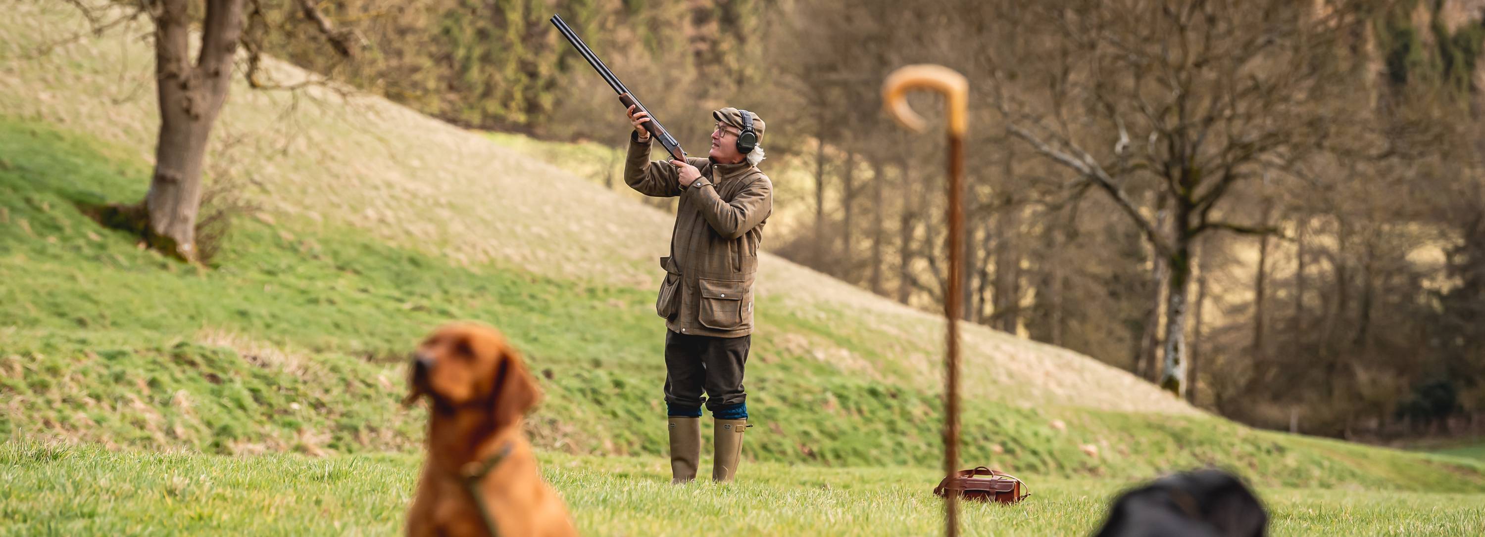 Simon Ward in brown jacket and green boots holds shotgun on hill