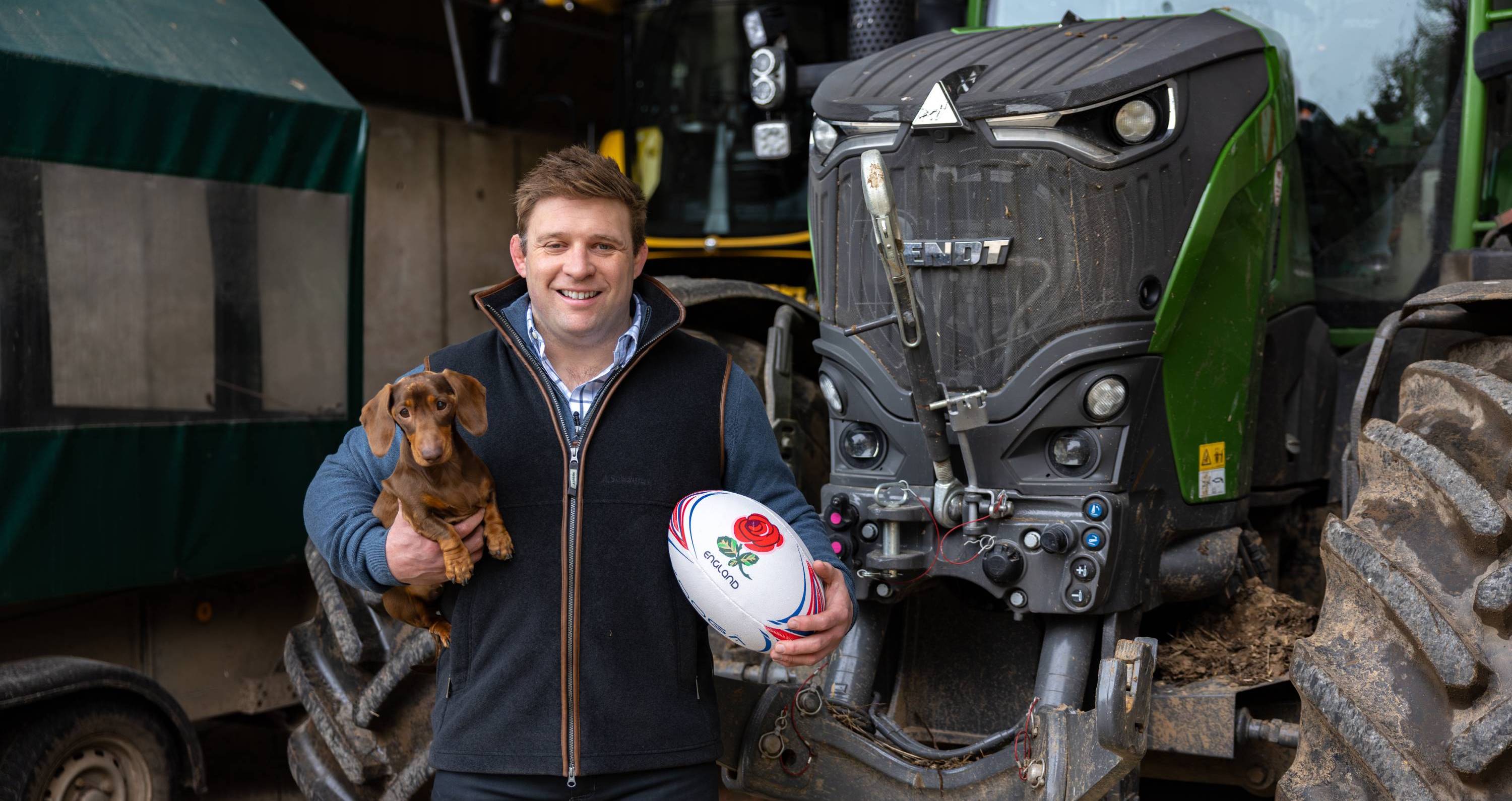 Tom Youngs holds white rugby ball with red rose and blue accents by green tractor