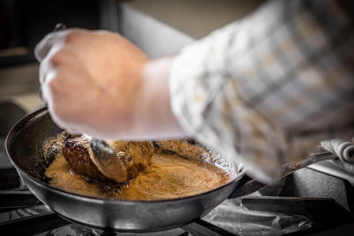 White gloved hand lifts seared venison steak from cast iron skillet with tongs