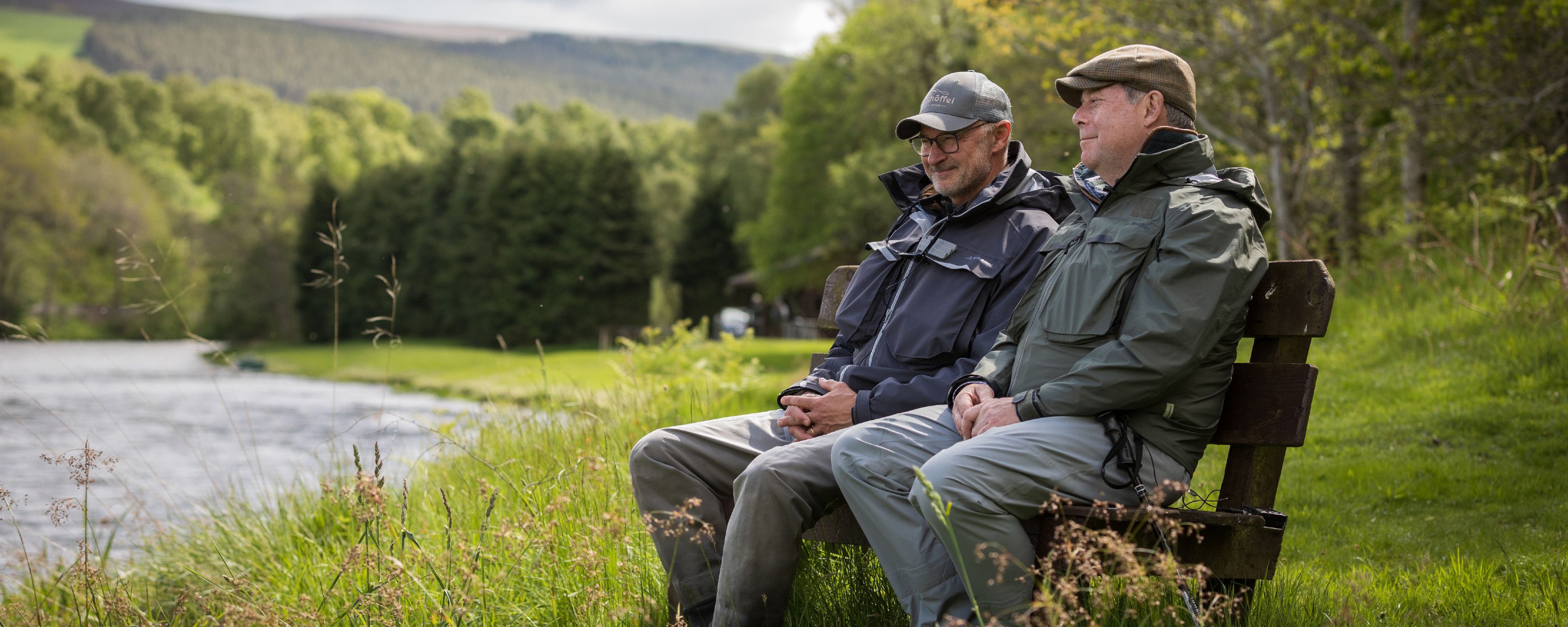 Two men in Schoffel attire on bench by river, celebrating 30 years