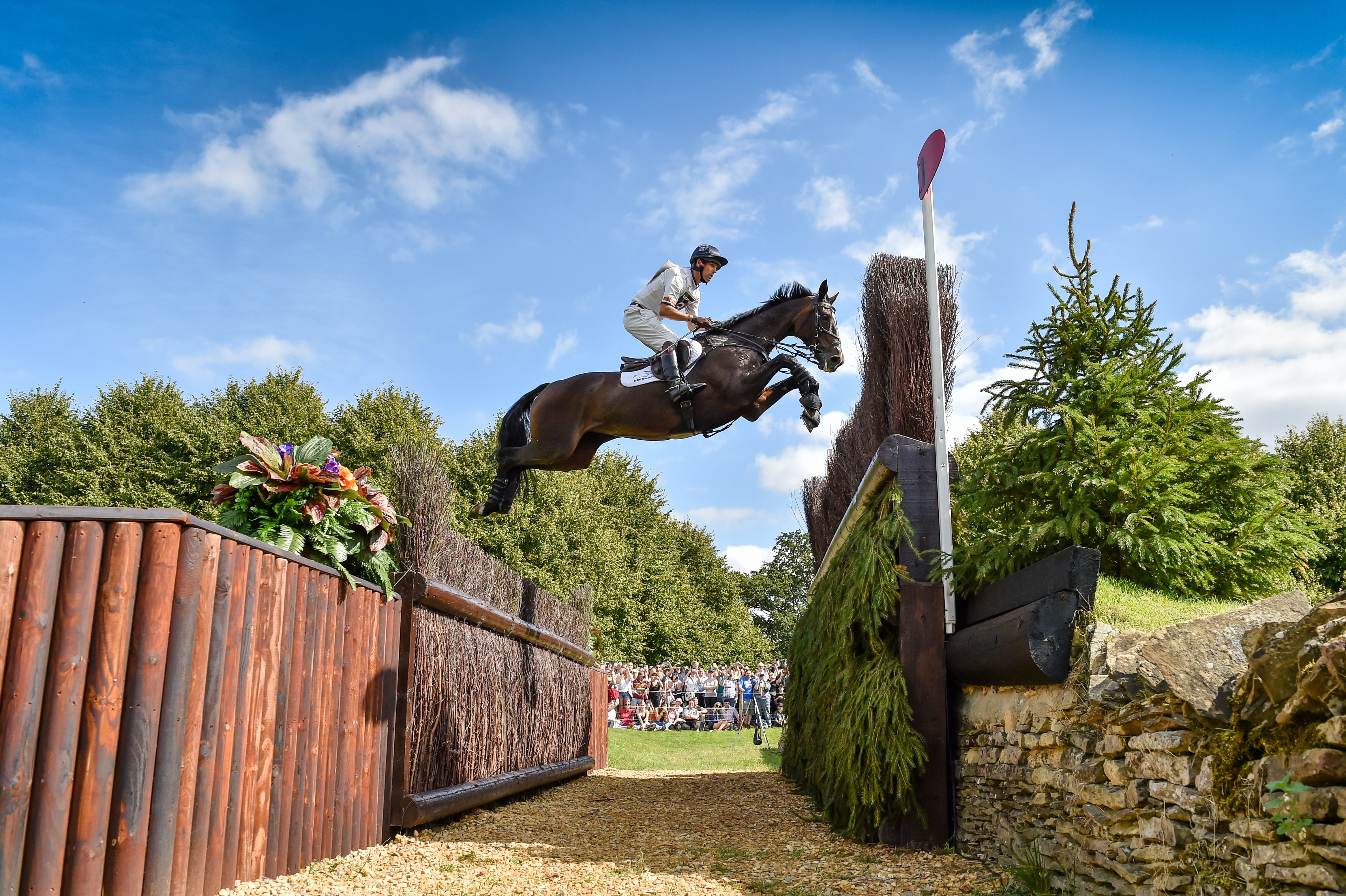 A dark brown horse with a rider in white equestrian attire jumps over an obstacle in an outdoor show jumping event under a blue sky with clouds.