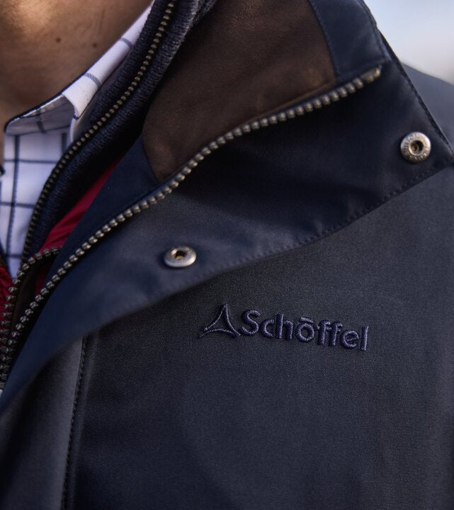 Close-up of a navy outdoor jacket showing the Schöffel embroidered logo, high collar and zip detail.