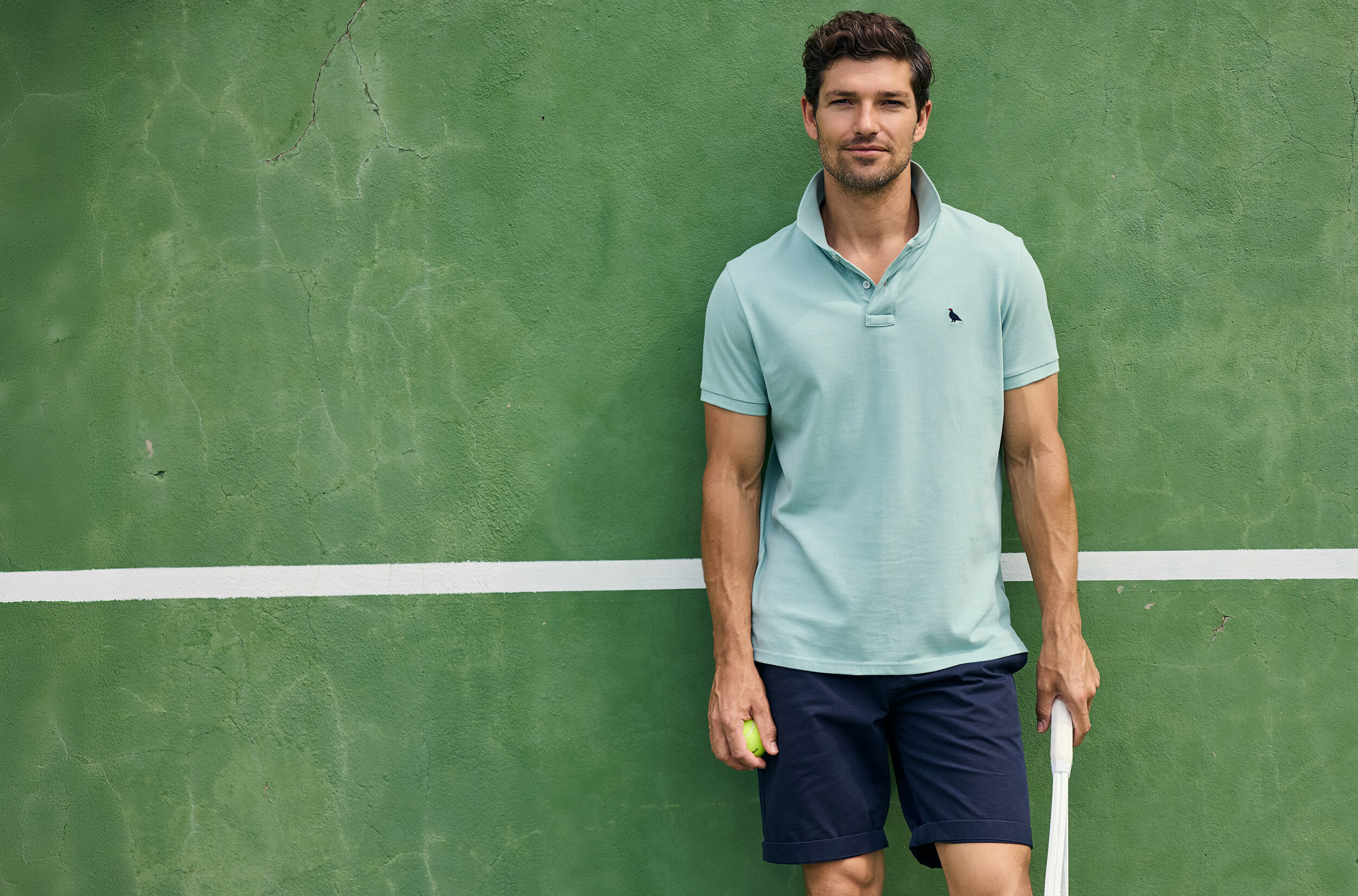 Man in light blue polo shirt and navy shorts holding a tennis racket on a green tennis court.