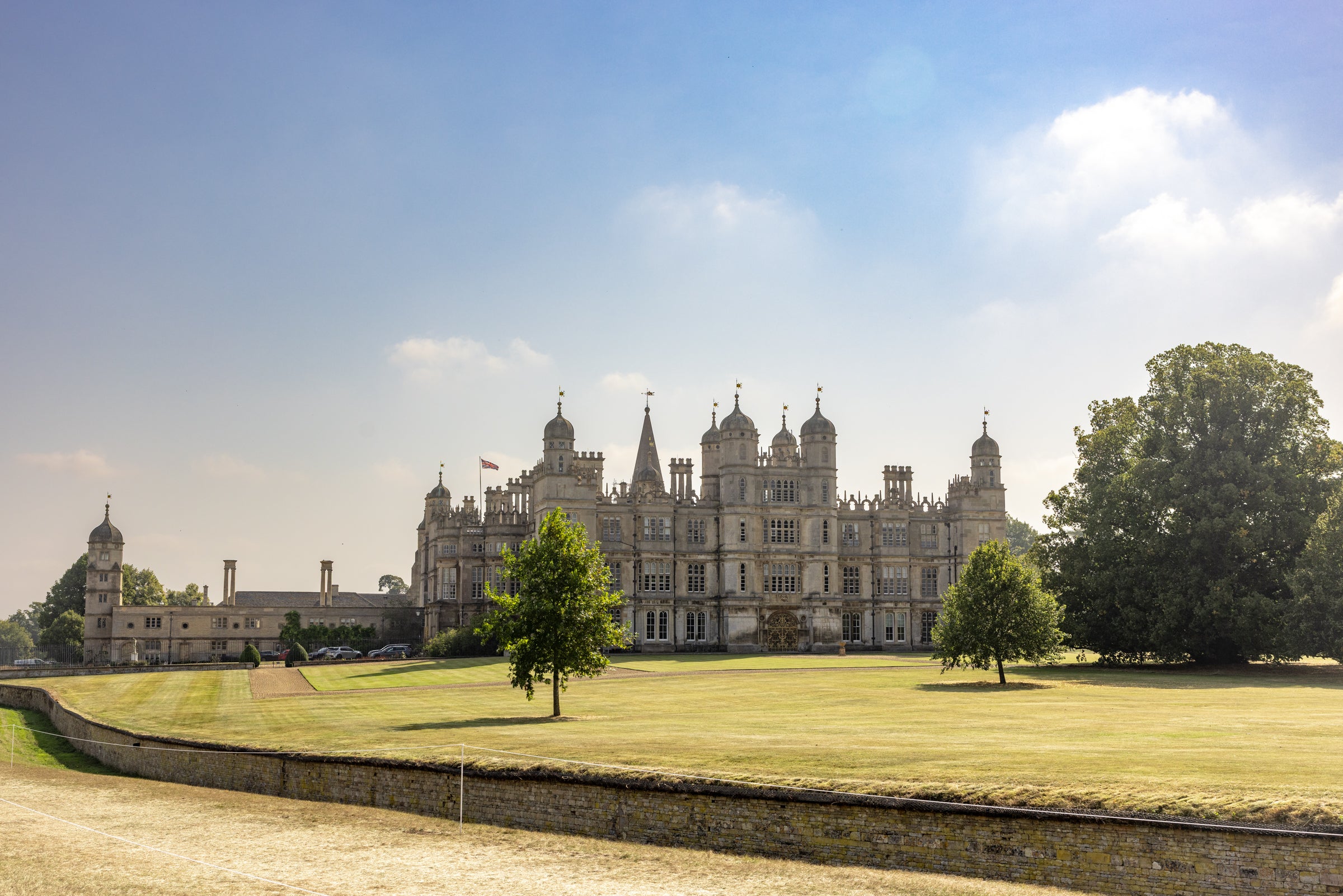 An picture of Burghley house featuring large manicured lawn under a blue sky with light clouds, with trees in the foreground.