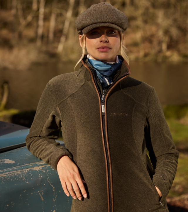 Person wearing a dark olive fleece jacket with zip fastening, leaning against a vehicle outdoors near water.