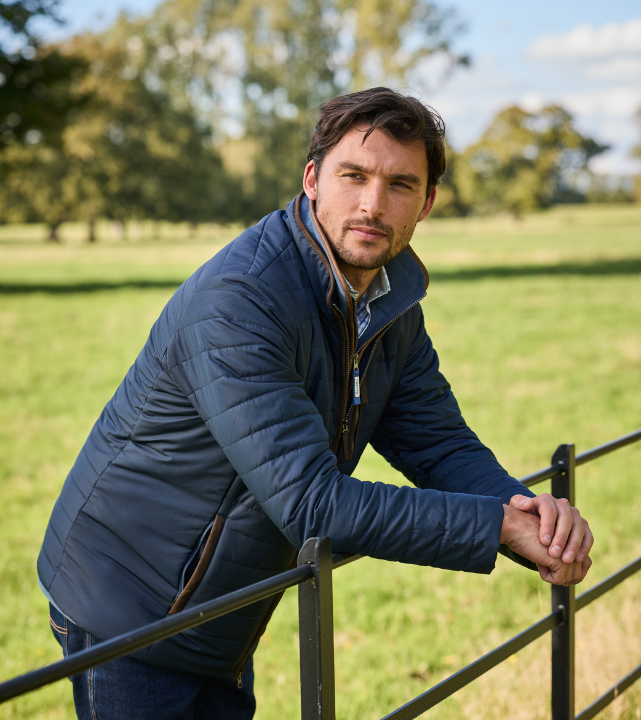 Person wearing a navy quilted jacket, leaning on a metal gate in a grassy countryside setting.