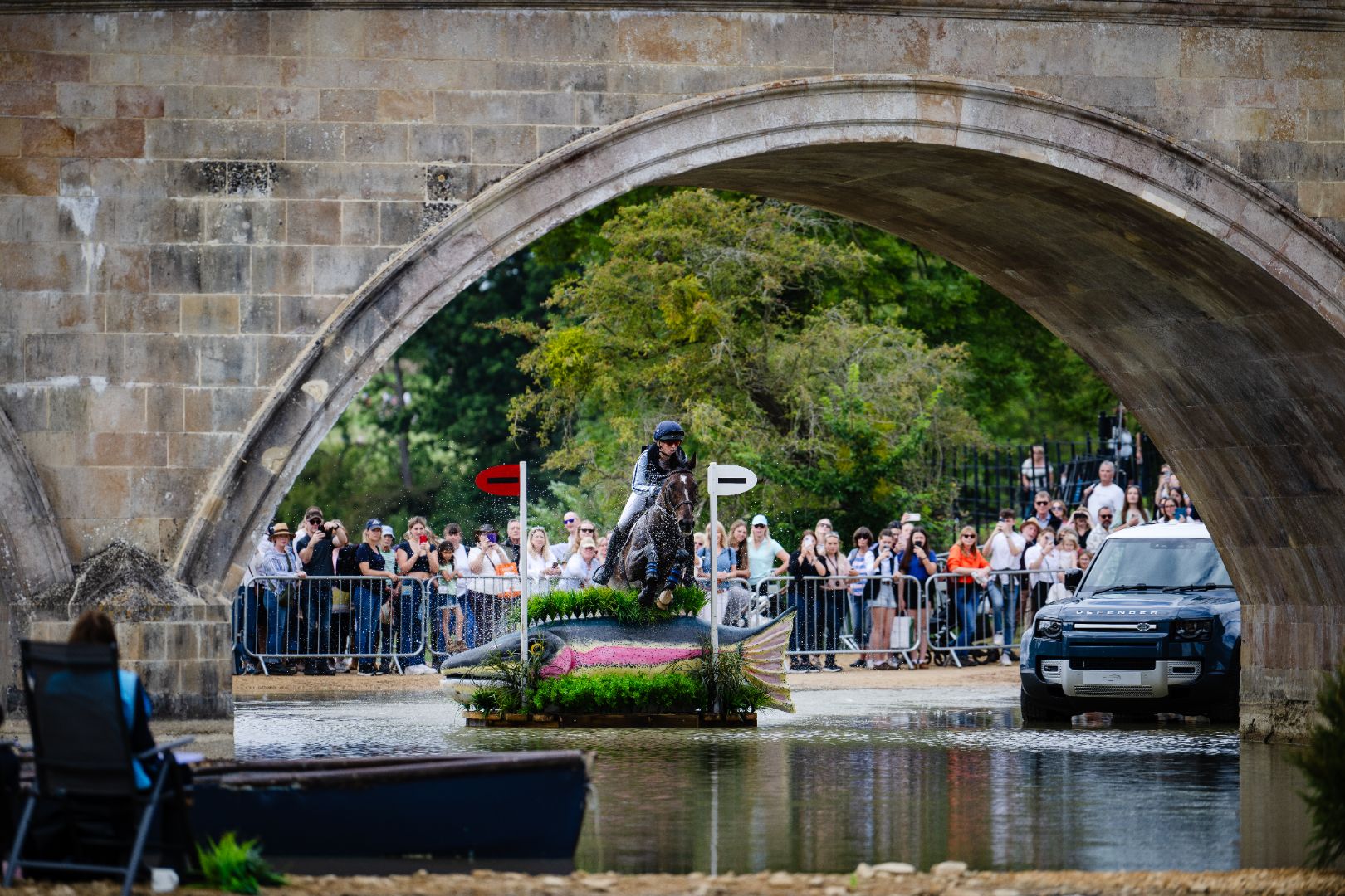 The beautiful setting for Burghley horse trials. Image shows a bridge over water with crowds of people watching a horse jumping an obstacle.