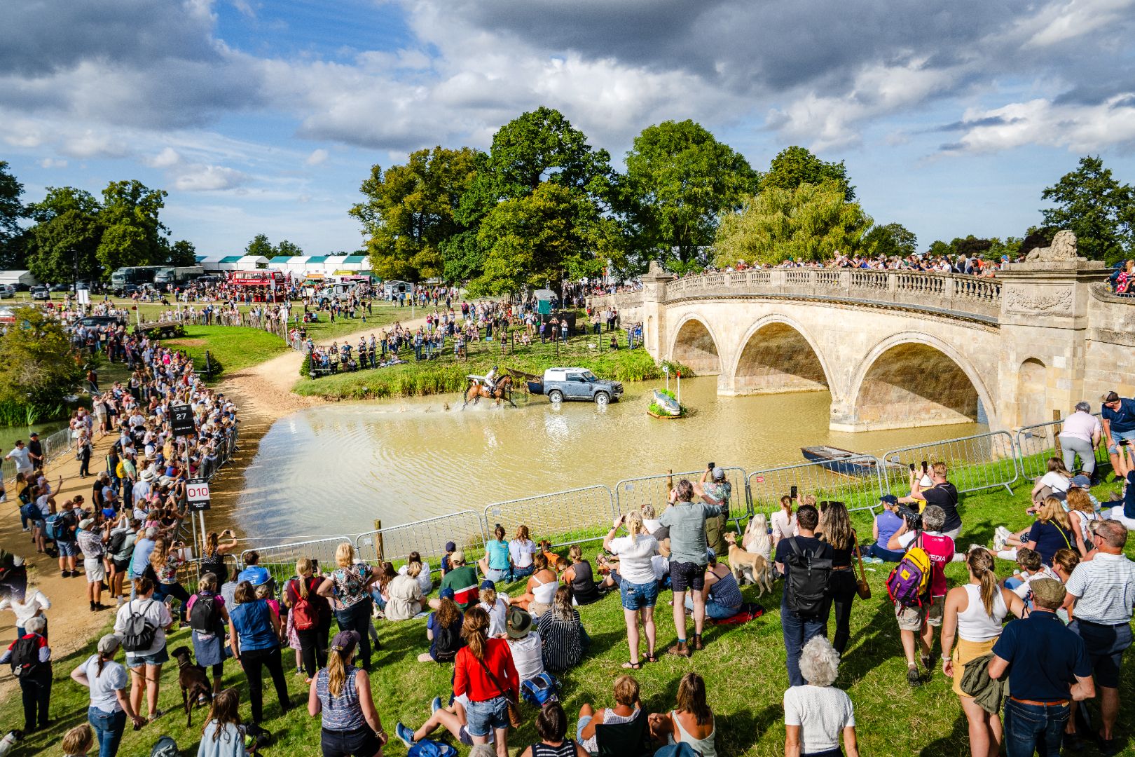 The beautiful setting for Burghley horse trials. Image shows a bridge over water with crowds of people watching on.