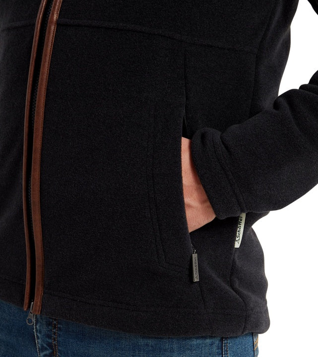 lose-up detail of a man's hand in the front pocket of a gunmetal grey fleece jacket, showing the tan leather-trim full-length zip and branded Schöffel zip pull, worn with blue jeans.