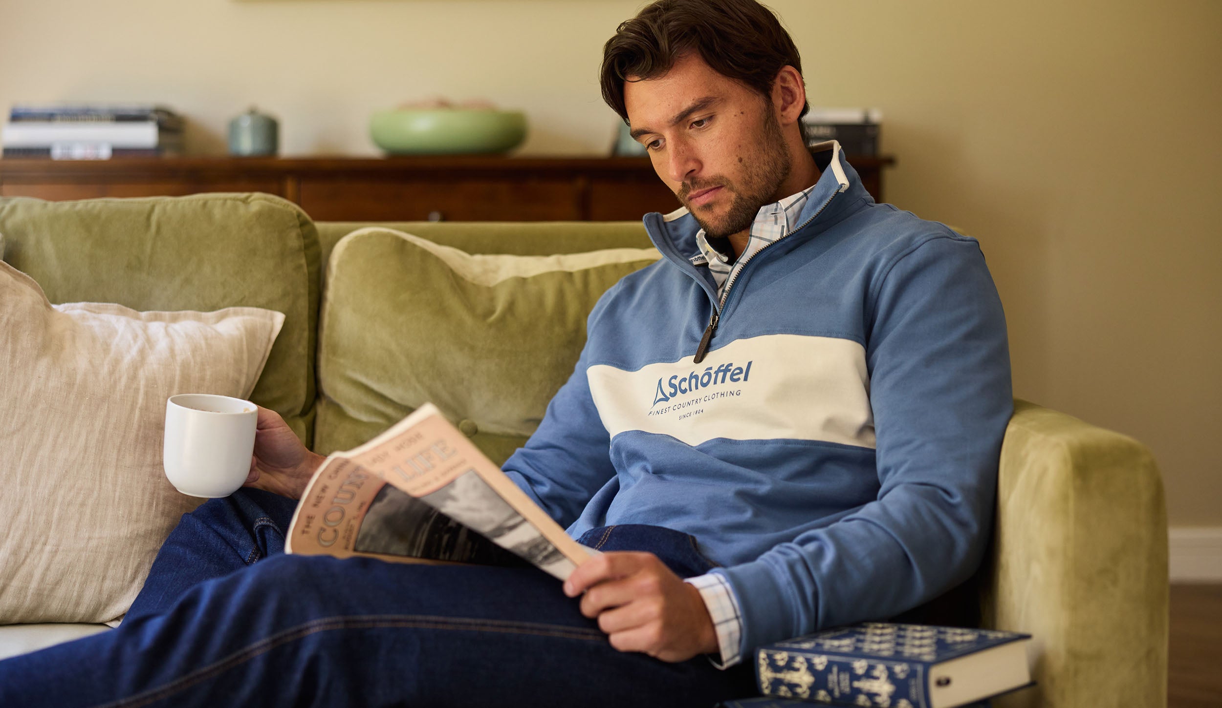 Man reading a book on a green couch wearing a blue sweatshirt with a logo.