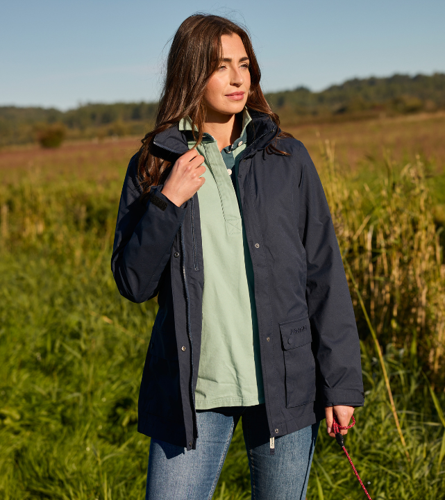 Person wearing a true navy outdoor jacket layered over a light green top, standing outdoors in grassy countryside.