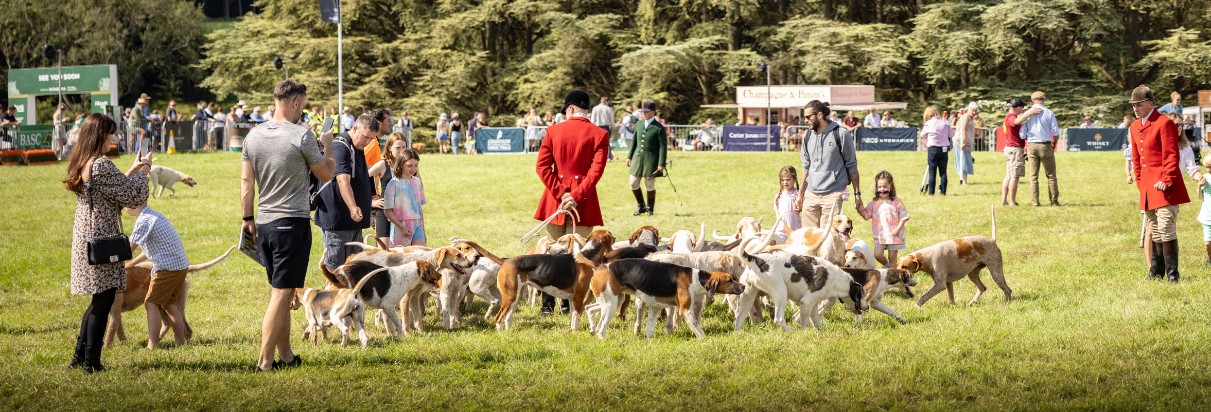 Visitors at The Game Fair interact with a pack of hounds, guided by traditionally dressed handlers, on a sunny field with vendor stalls in the background.