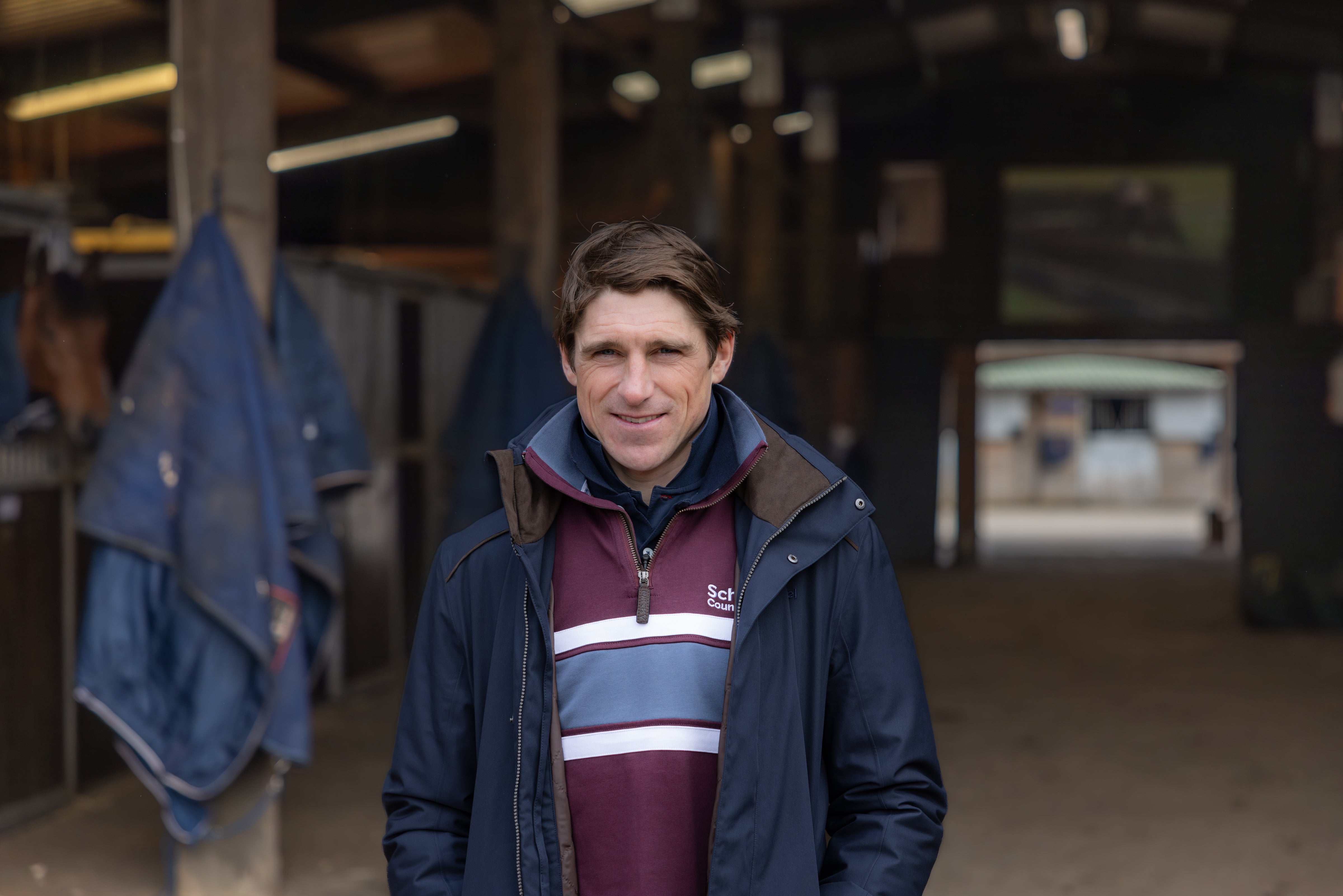 Harry Skelton in a navy jacket and maroon quarter zip sweatshirt smiling in a stable, with horse blankets and stalls in the background