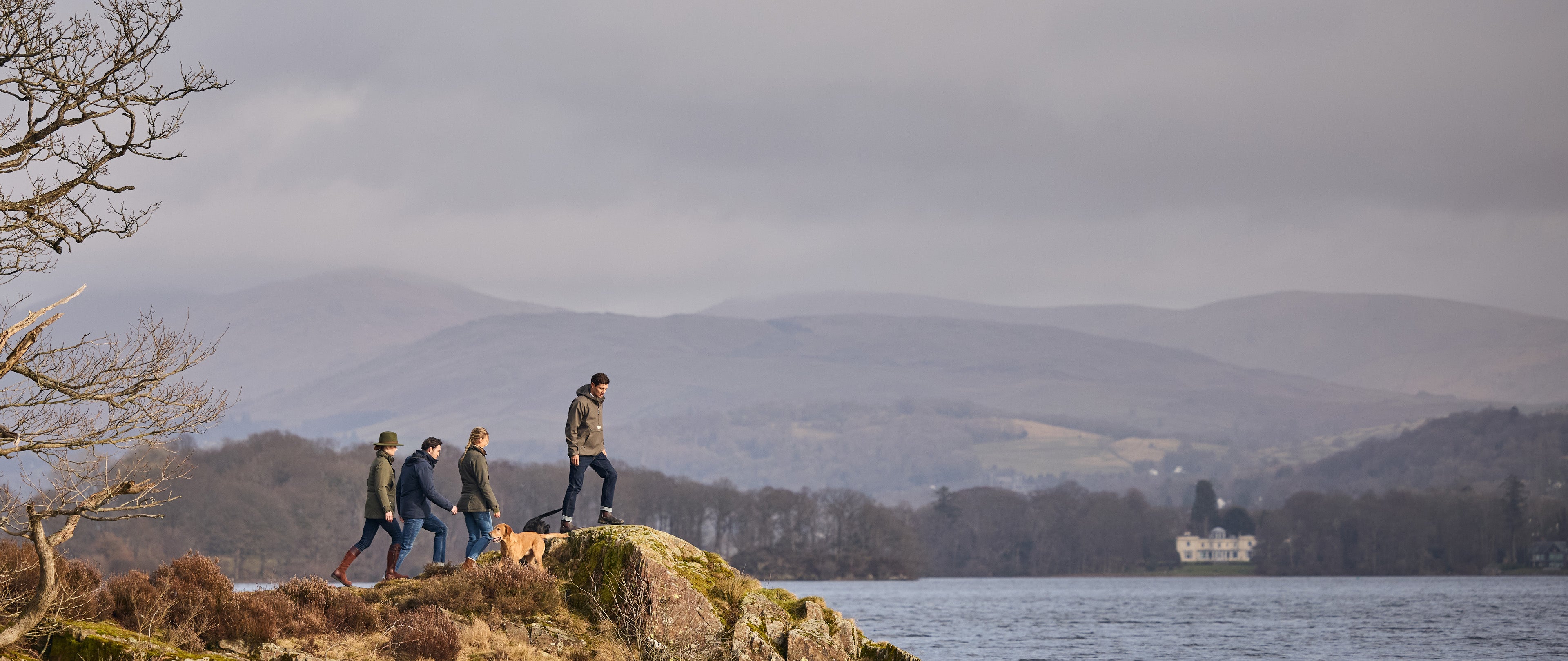 Four people walking a dog on a rocky terrain by a body of water with mountains in the background.