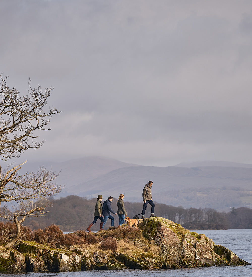 Four people walking a dog on a rocky terrain by a body of water with mountains in the background.