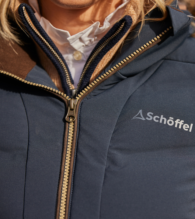 Close-up of the chest area of a carbon navy down coat, featuring a large brass-toothed zip with a brown leather zip trim and fleece-lined collar. The silver Schöffel logo is printed on the chest. A white shirt is visible underneath.