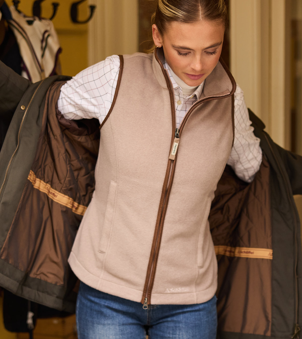 A woman wearing a light beige zip-up fleece gilet over a white checkered shirt and blue jeans, holding open a dark brown quilted jacket to show the interior lining.