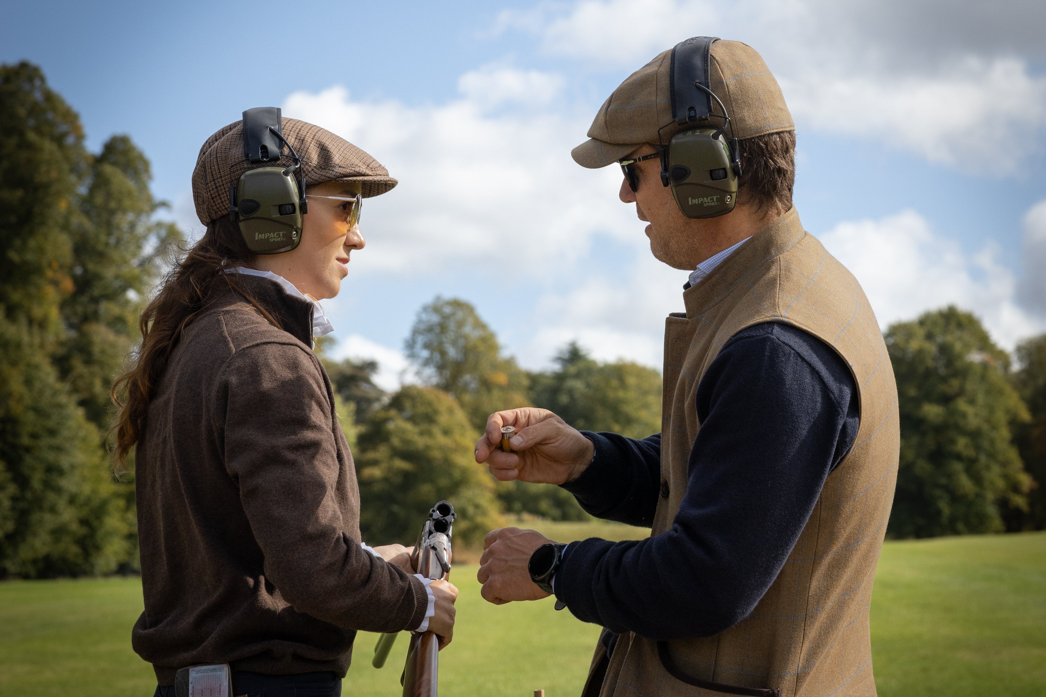 Two men in outdoor attire with guns and ear protection on a grassy field.
