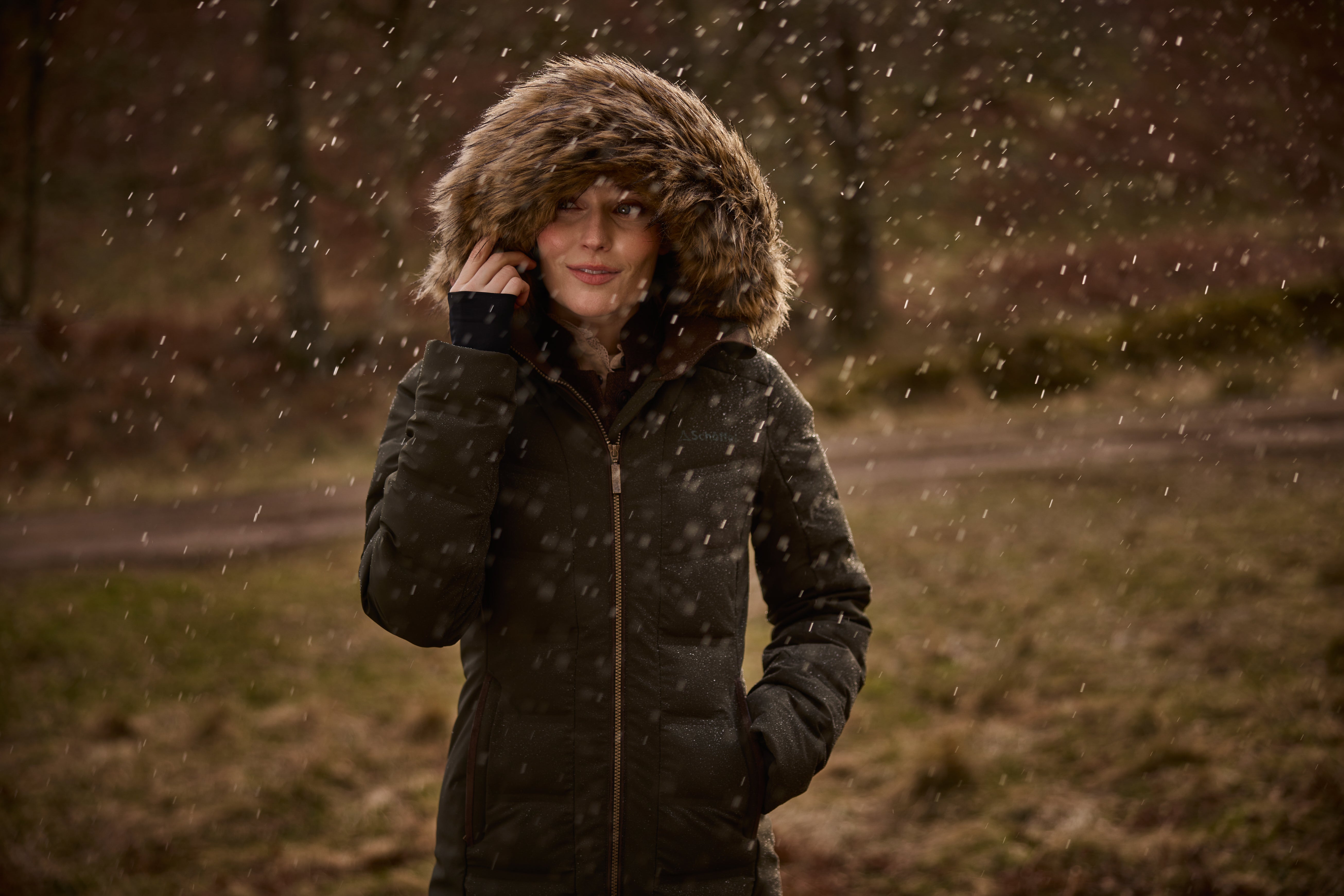 A woman wearing a dark green winter parka with a large brown faux-fur trimmed hood stands outdoors in falling rain