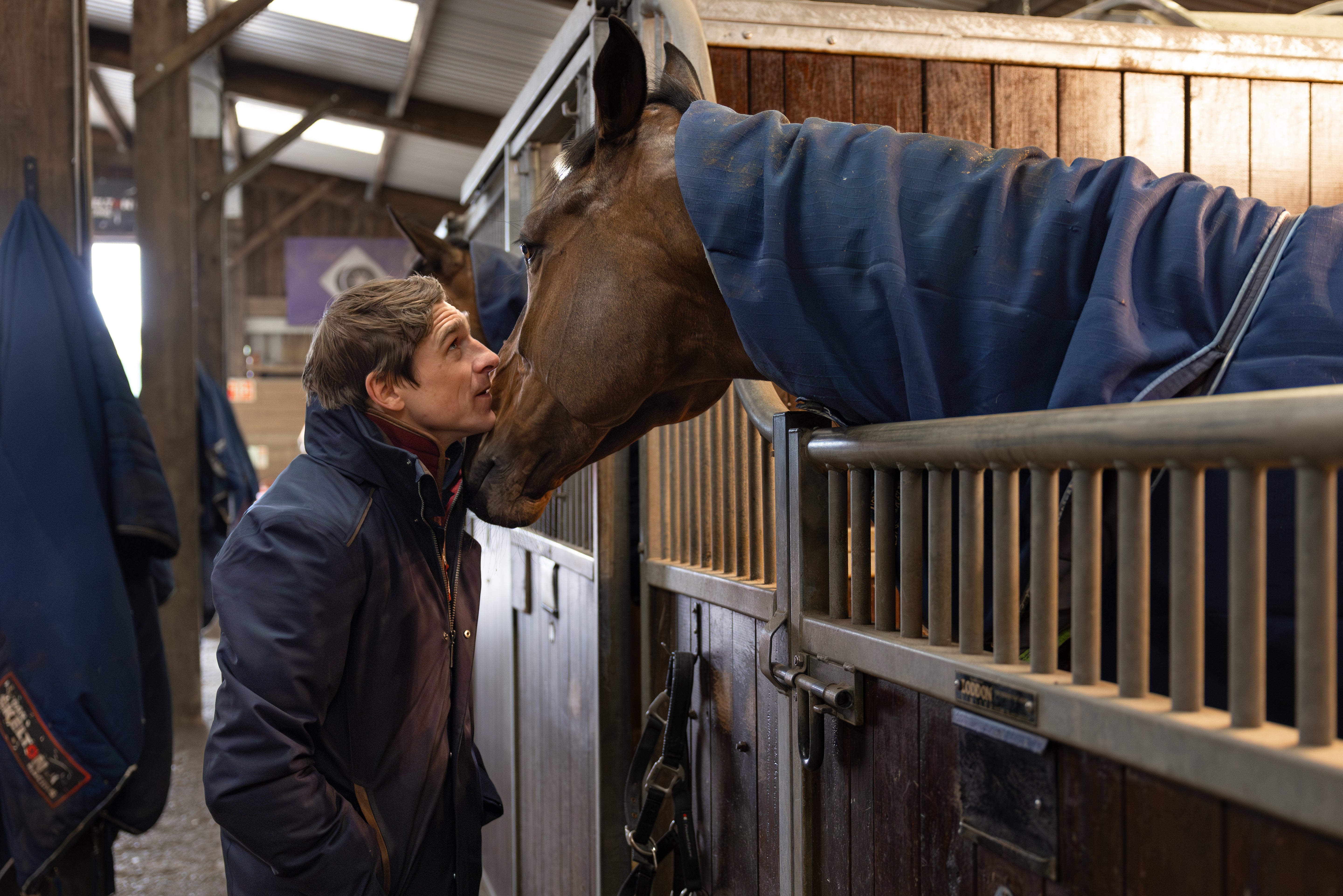Harry Skelton shares a close moment with horse Protektorat in the stable, touching noses as the horse leans over the stall door, wrapped in a navy blanket