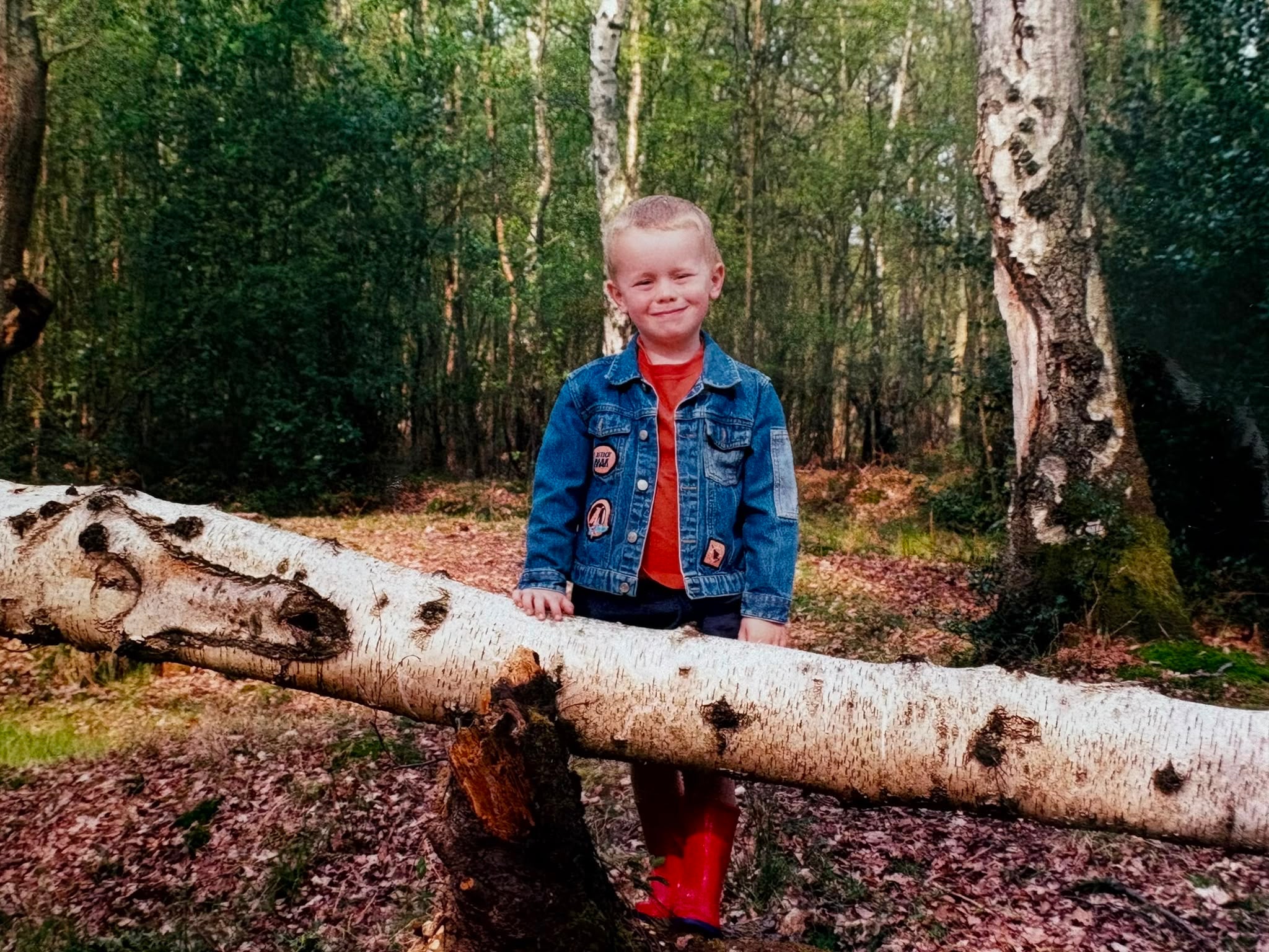 A young Tom Hilder standing on a fallen birch log in a forest, wearing a blue denim jacket with decorative patches over an orange shirt and red rain boots.