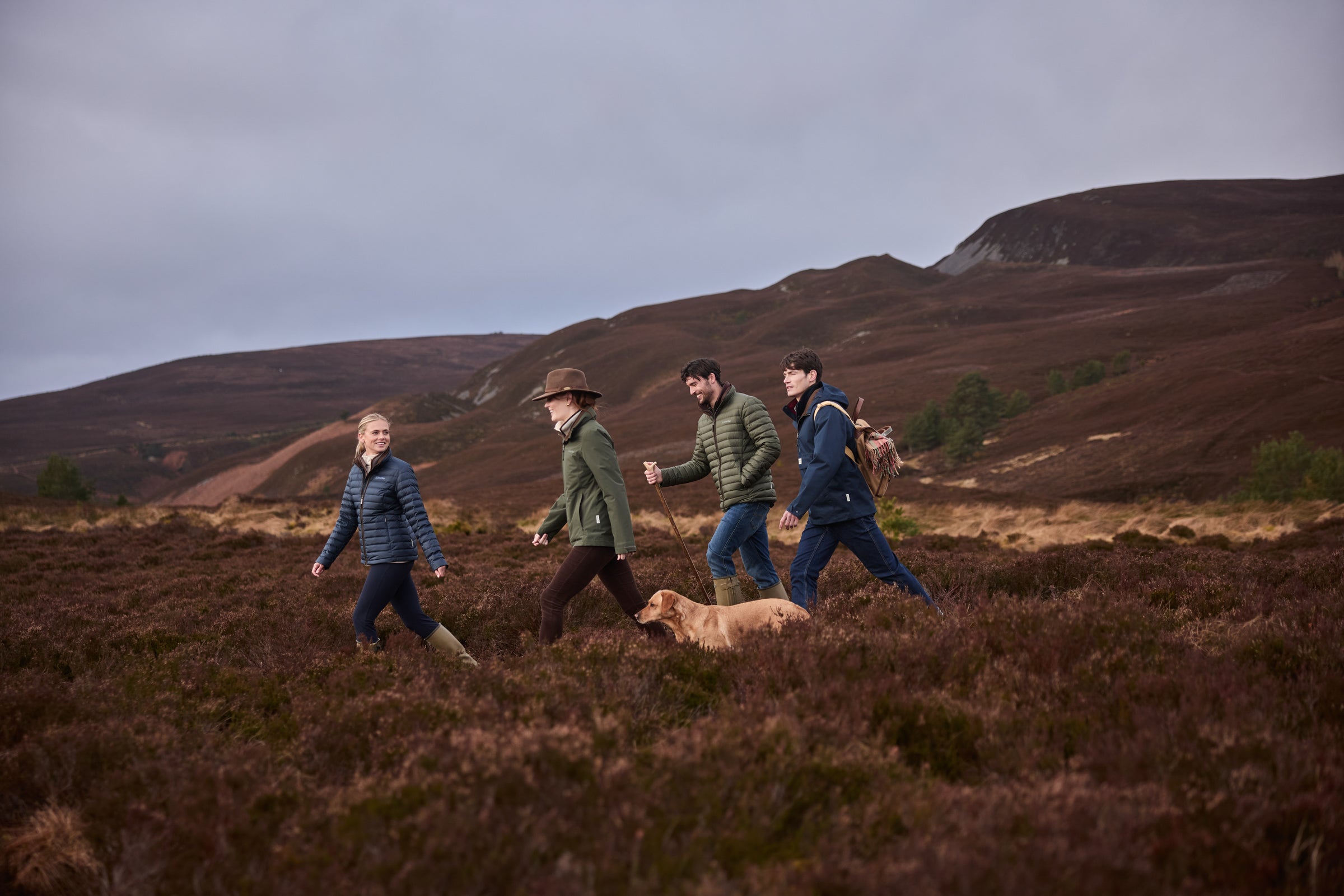 Four people walking across a moorland landscape with rolling hills in the background under an overcast sky.