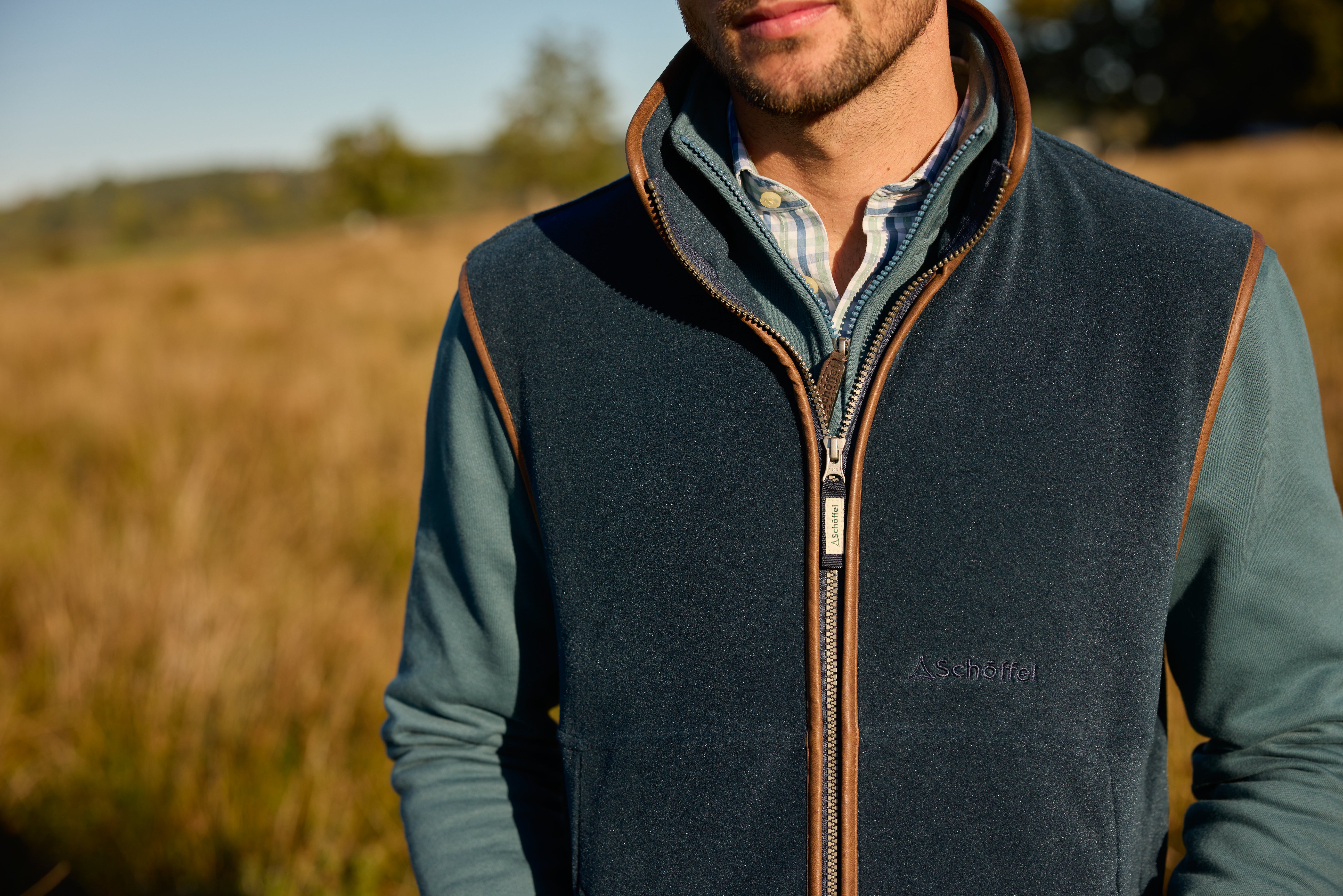 Schöffel men's fleece gilet in navy with tan leather trim, close-up on model layered over a sage green fleece and checked shirt, worn outdoors in a countryside field