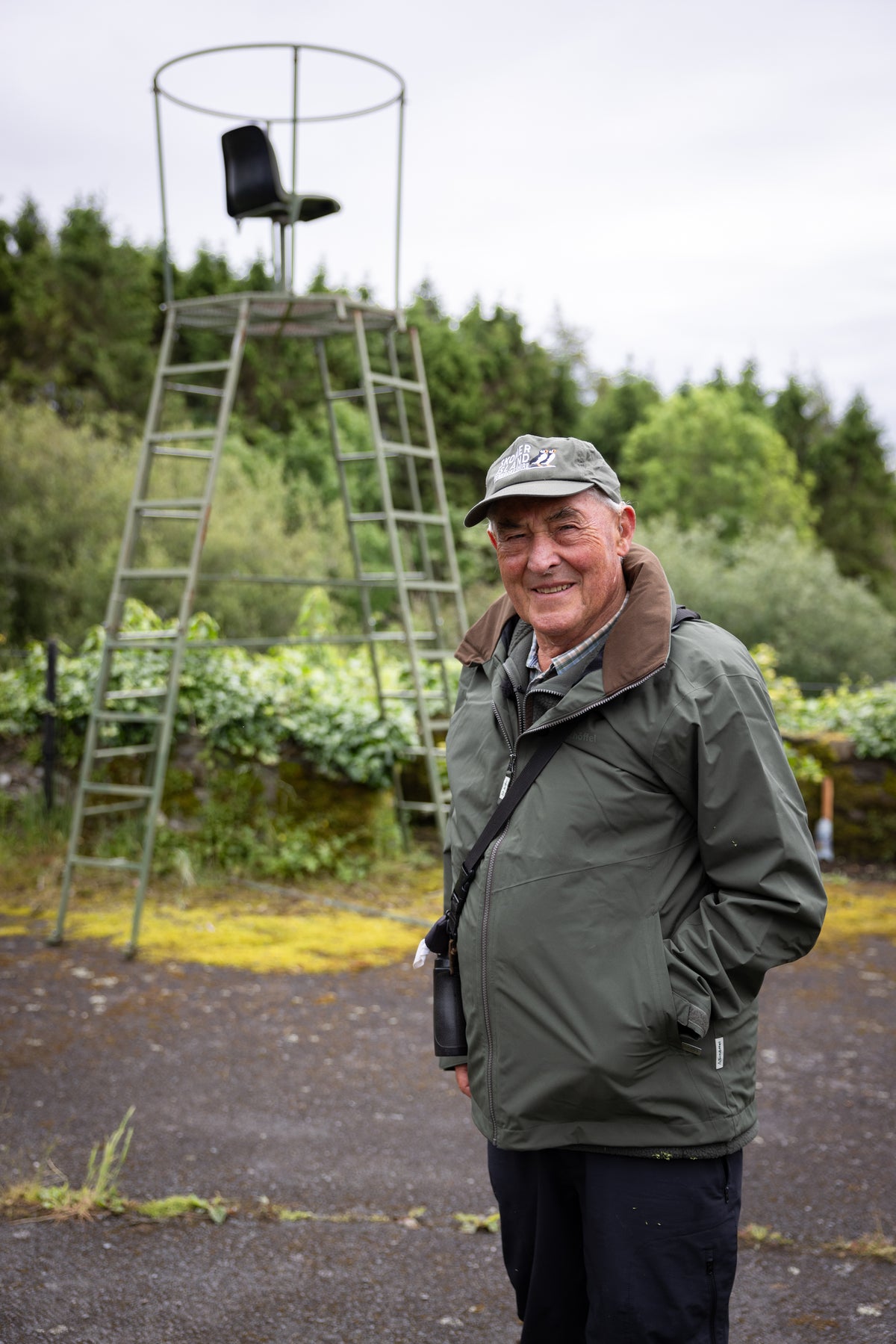 Smiling man in a green jacket and grey cap standing by a forest and watchtower.