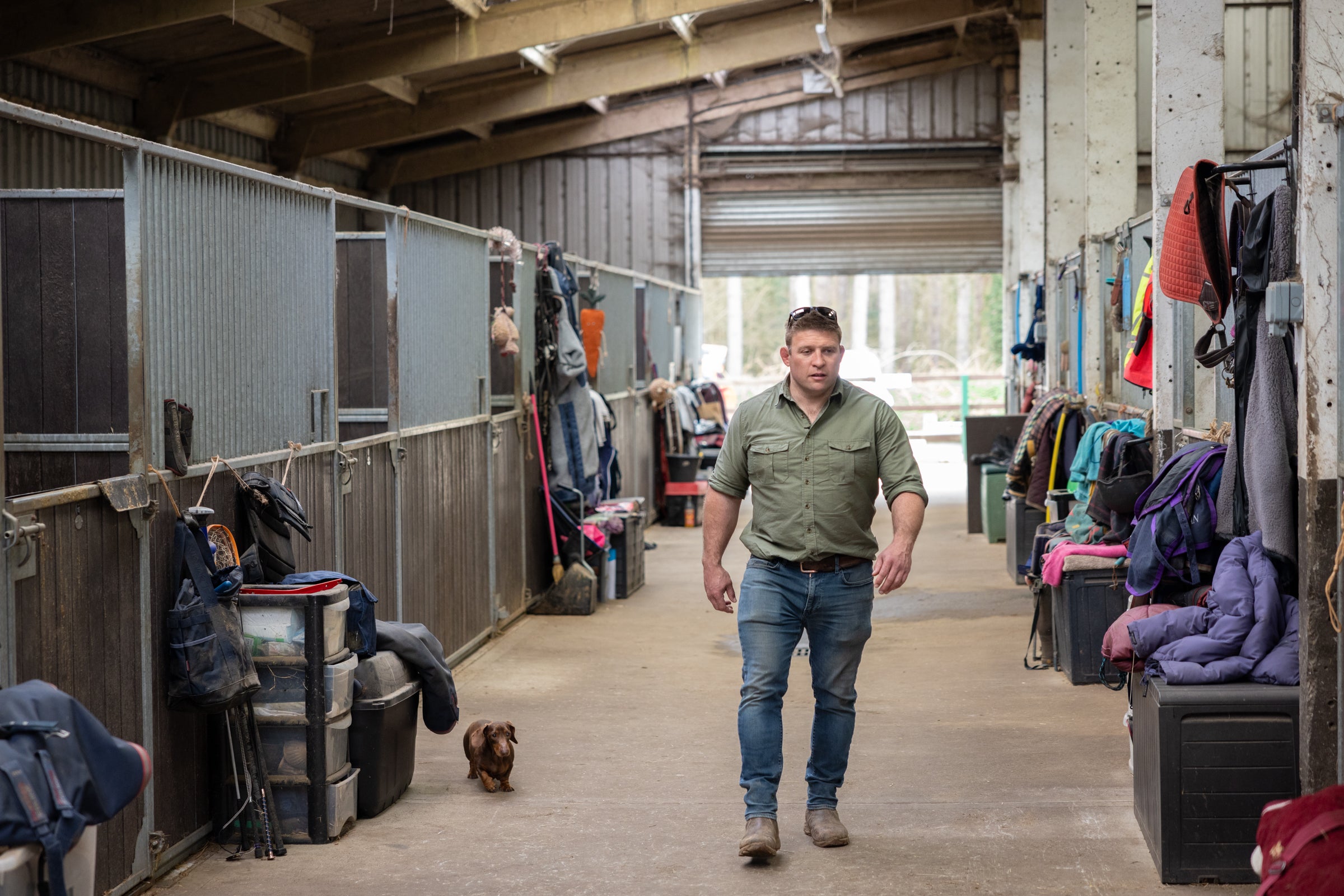 Man walking through a stable with horses and equipment around
