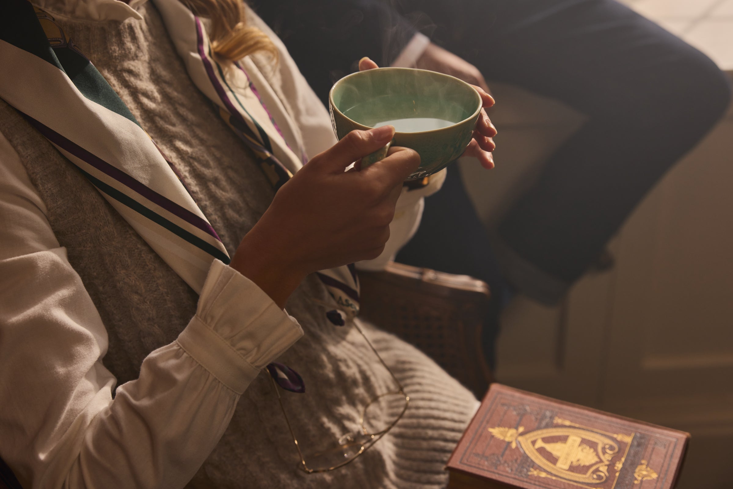 A close-up lifestyle shot of a person holding a green ceramic teacup, with a vintage book.
