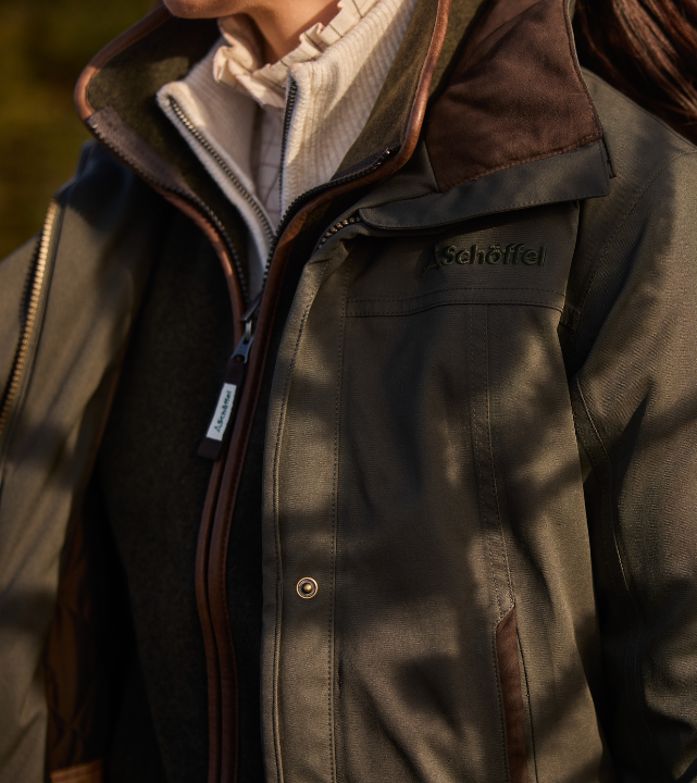 Close-up of the front of a dark woodland green country coat with a zip fastening, brown leather detailing on the collar and shoulders, a press-stud chest pocket, and embossed Schöffel branding. A cream knitted gilet and white shirt are visible underneath.