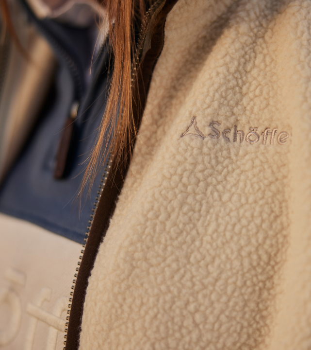 Close-up of the chest of an ecru textured fleece gilet, showing the tonal Schöffel logo embossed into the fabric. A dark brown leather-effect zip trim runs along the front opening, with a glimpse of a navy layer visible underneath.