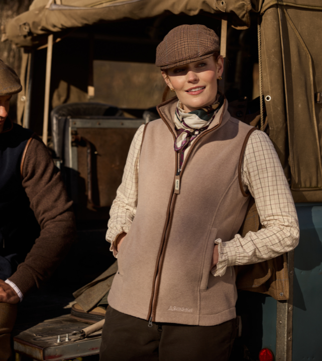 A person stands beside a vintage off‑road vehicle, wearing a checked shirt, a light brown gilet, a patterned neck scarf and a brown tweed flat cap. Their hands are in their gilet pockets, and another person in outdoor clothing is partially visible to the left. The scene appears to be set outdoors in warm, late‑afternoon light.