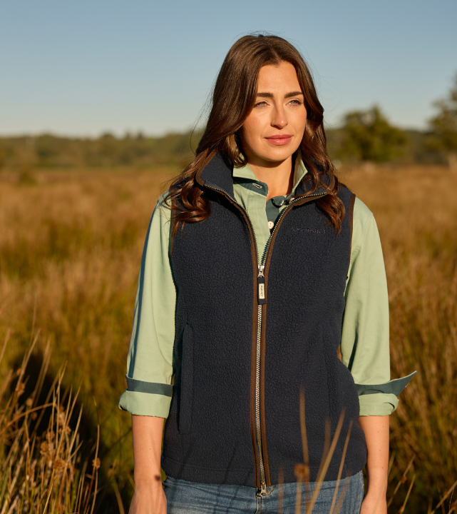 Woman wearing a navy fleece gilet over a light green shirt, standing in a grassy countryside landscape on a sunny day.