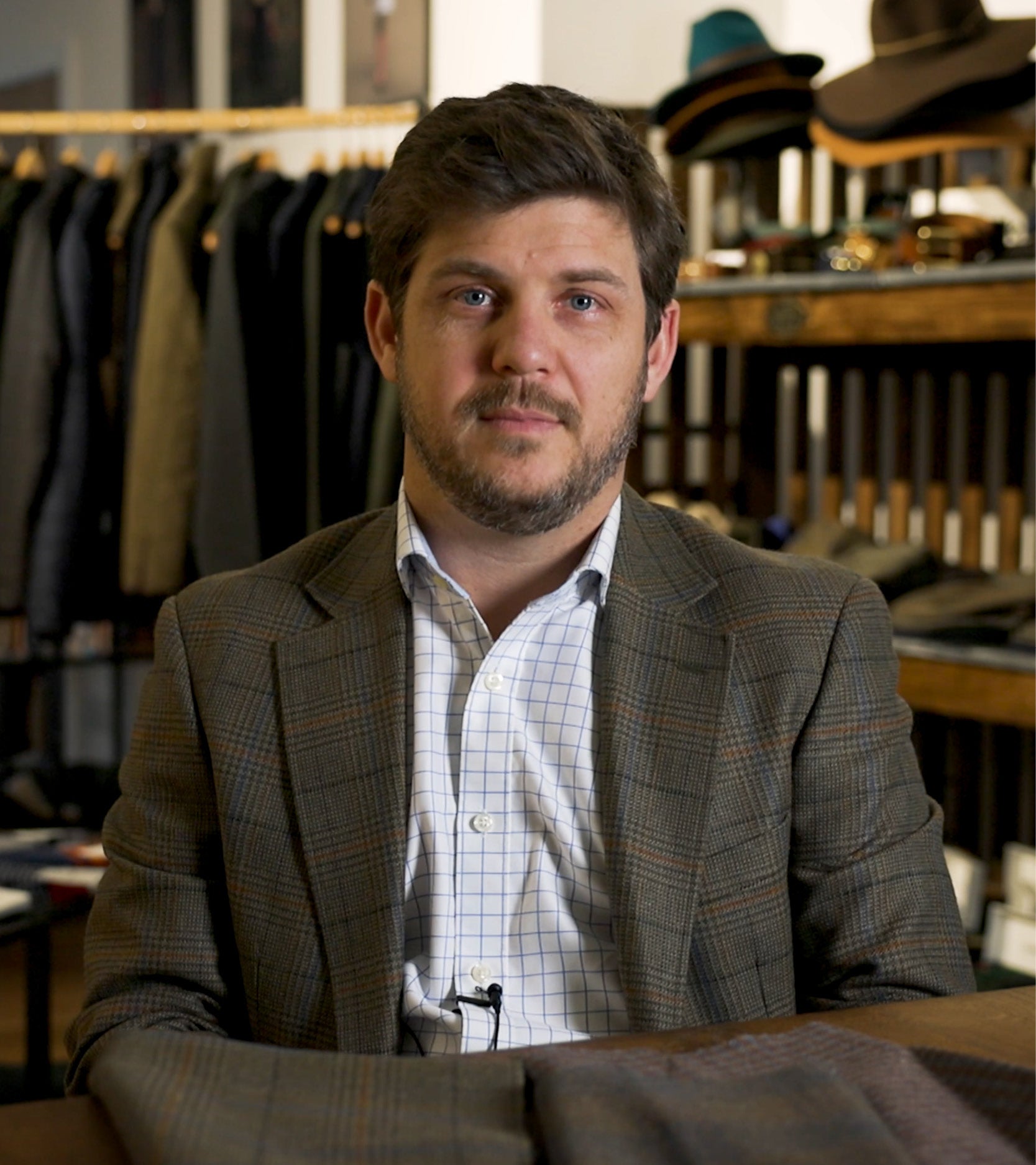 A man wearing a white shirt and tweed blazer. He is sitting down in a show room with other tweed clothing.