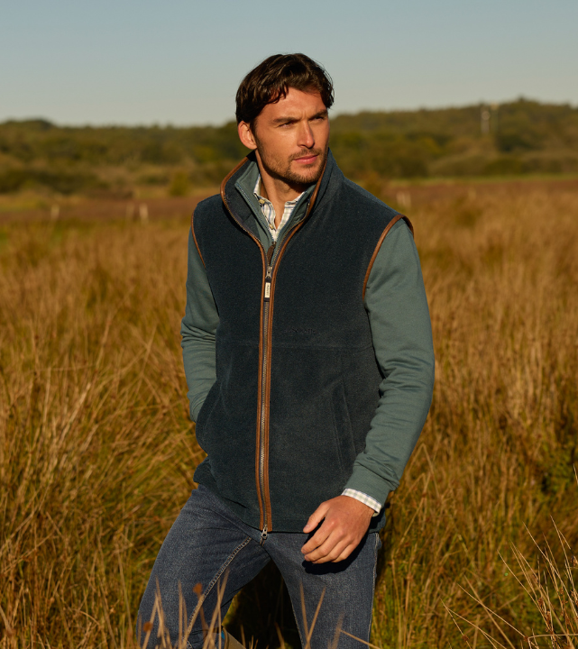 A man standing in a sunlit grassy field, wearing a navy fleece gilet over a long-sleeved top and shirt, looking off into the distance.