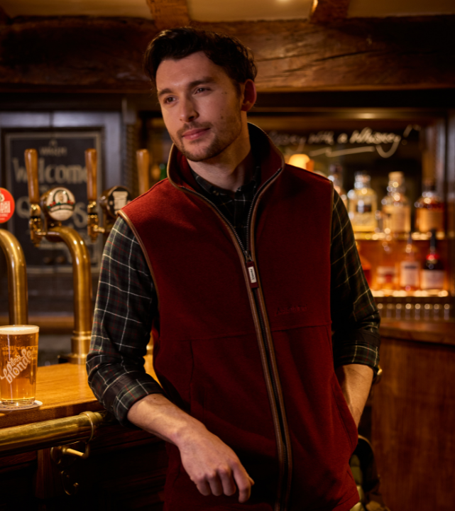 Person wearing a claret fleece gilet layered over a shirt, standing indoors in a pub setting.