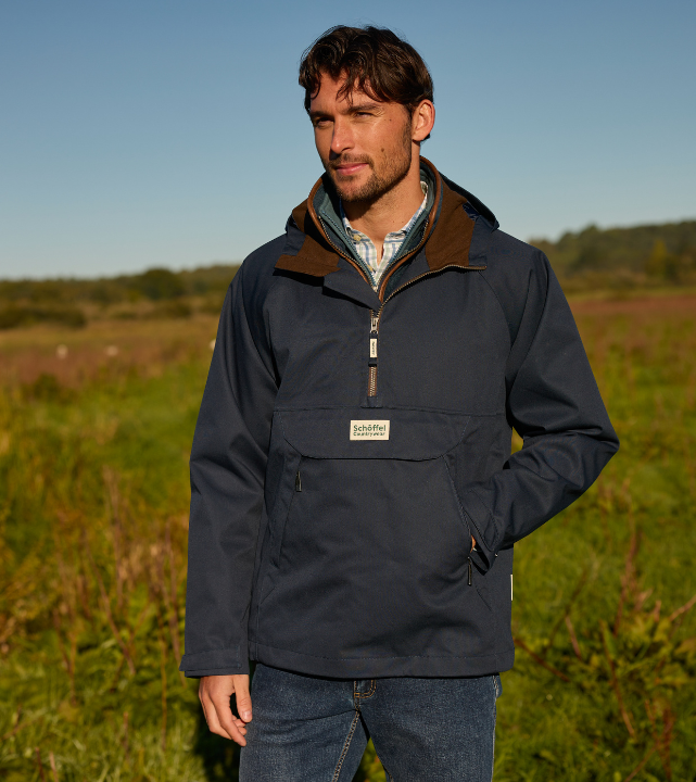 A man standing outdoors in a sunny rural landscape, wearing a navy smock-style jacket over a shirt, with one hand in his pocket, looking slightly to the side.