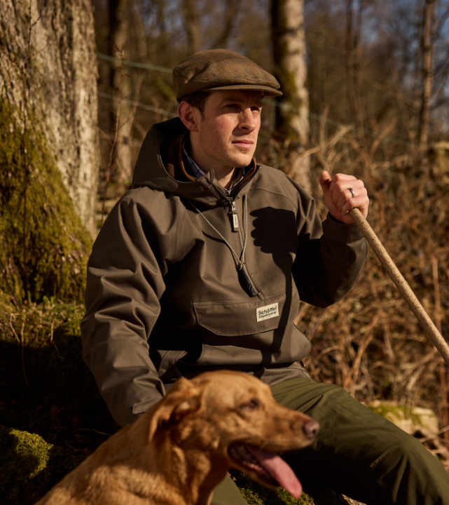 Person wearing an olive outdoor jacket and flat cap, sitting outdoors in woodland with a dog.