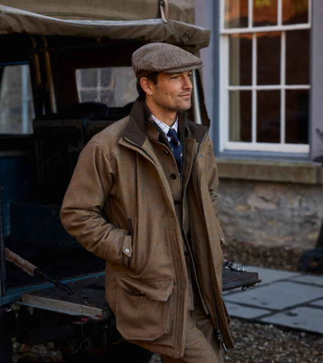 Person wearing a corry tweed country coat and flat cap, standing outdoors beside a vehicle.