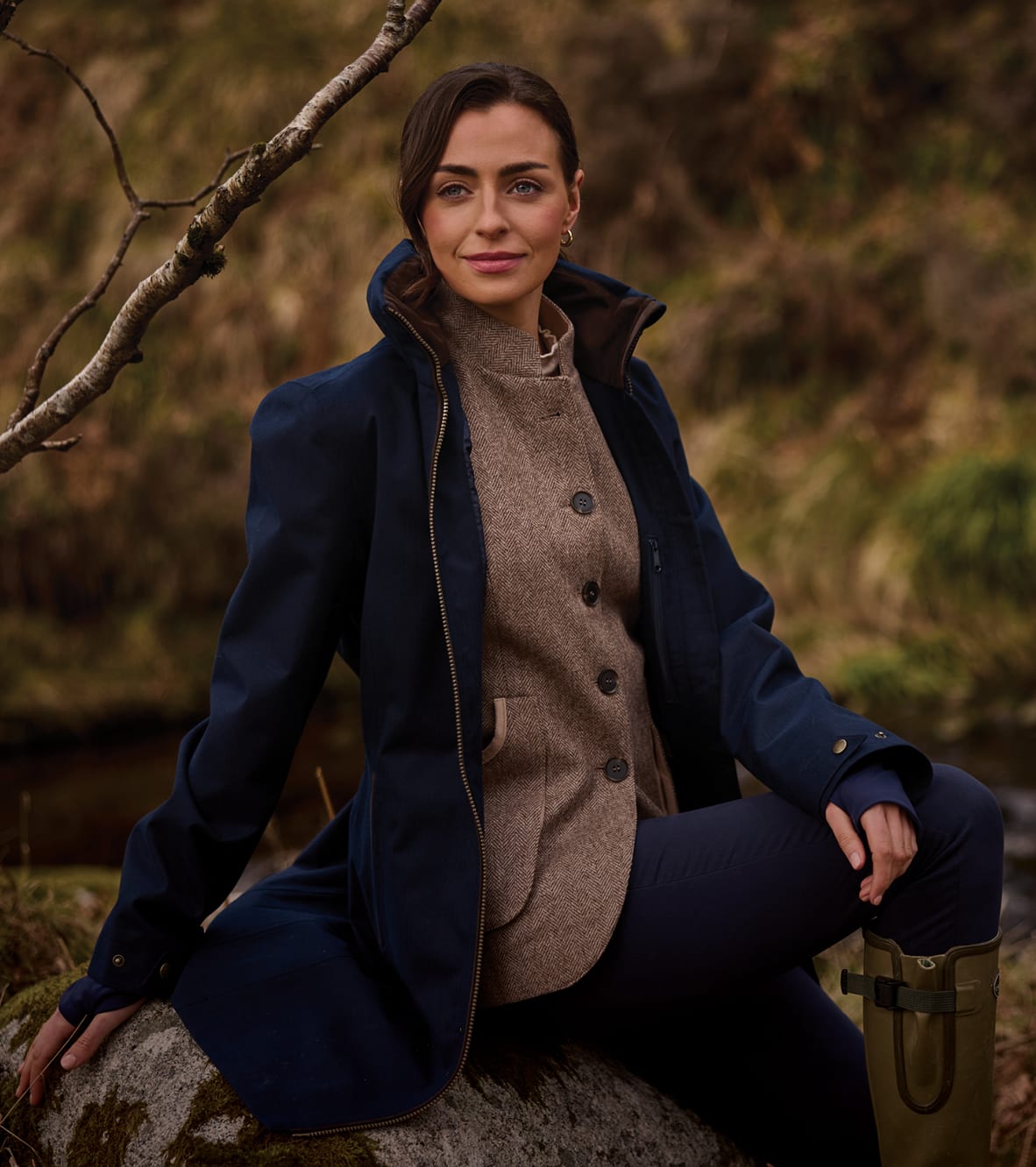 A woman sits outdoors on a rock wearing a navy blue coat over a brown tweed waistcoat.
