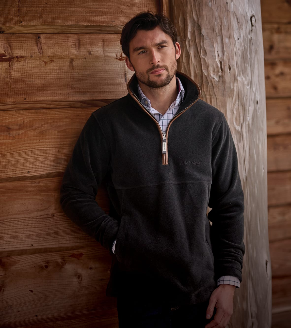 A man wearing a dark grey quarter-zip fleece standing against a rustic wooden wall.