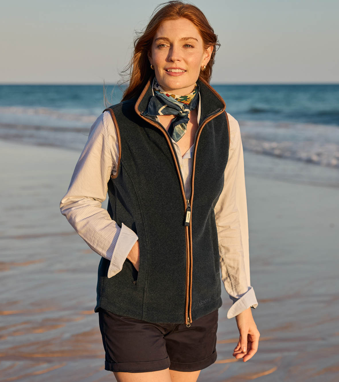 Woman standing on a beach wearing a dark vest and scarf with ocean waves in the background
