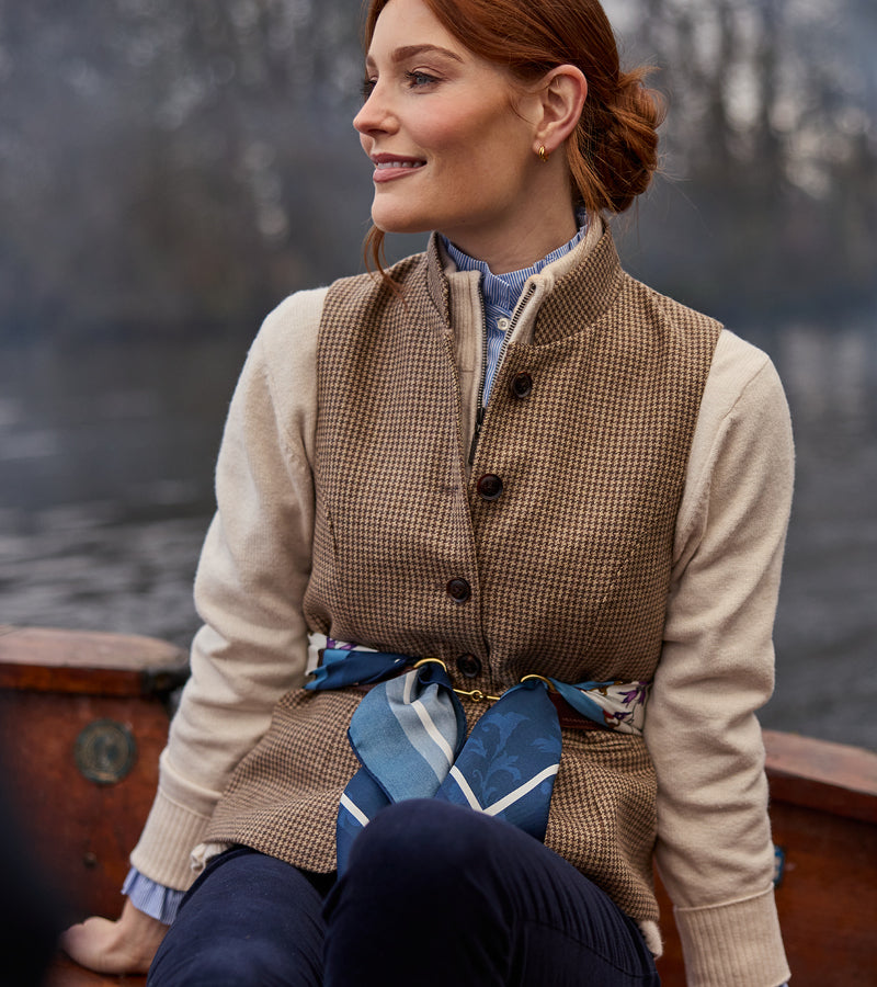 Woman wearing a brown checked waistcoat over a beige sweatshirt with a blue scarf, sitting outdoors.