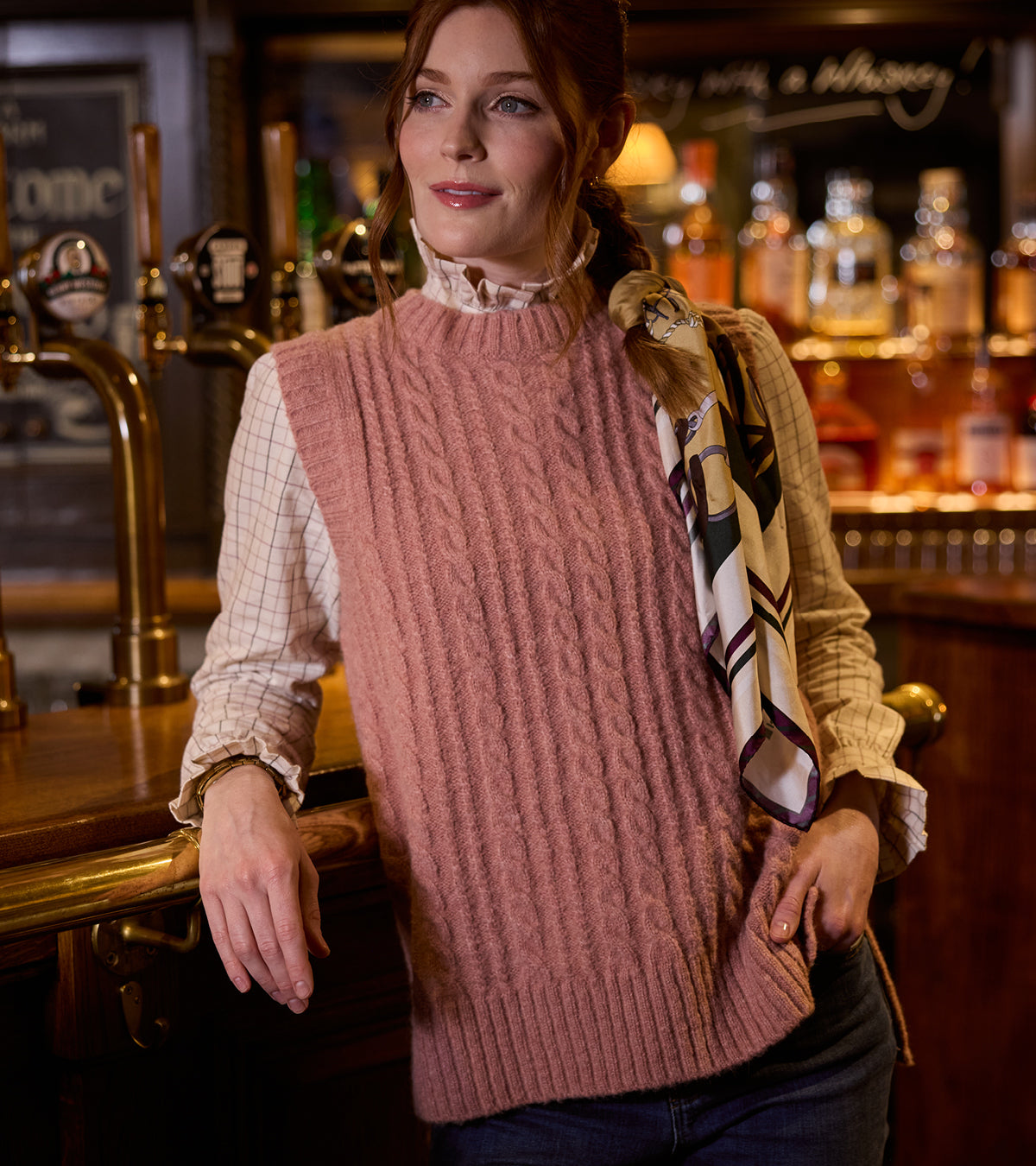 Woman with a silk scarf in her hair wearing a pink knitted vest in a bar.