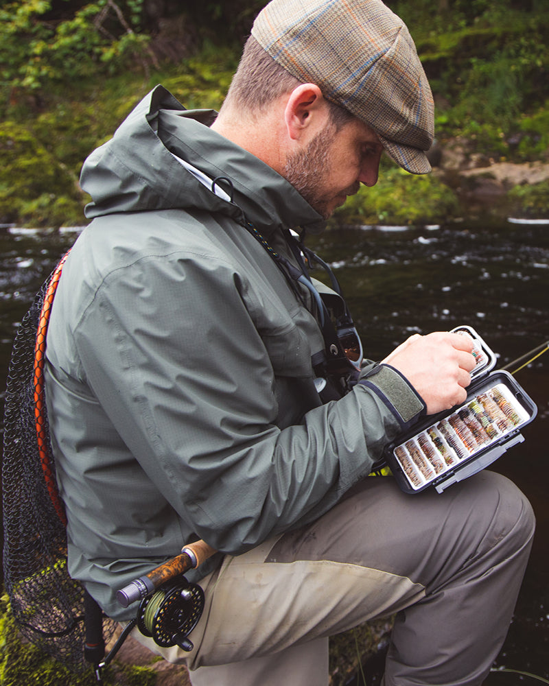 Man in fishing gear by a river, holding a fly fishing box with flies.